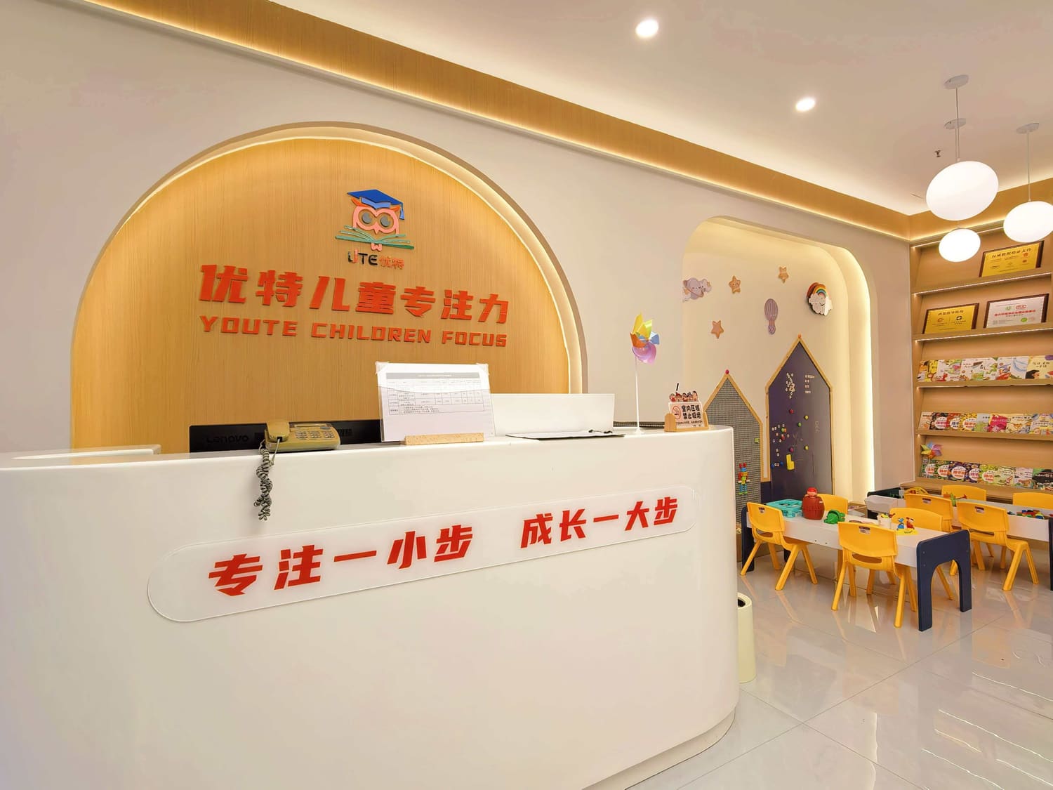 校区前台实景：木饰面 logo 墙与「专注一小步 成长一大步」标语，右侧是儿童桌椅与书架